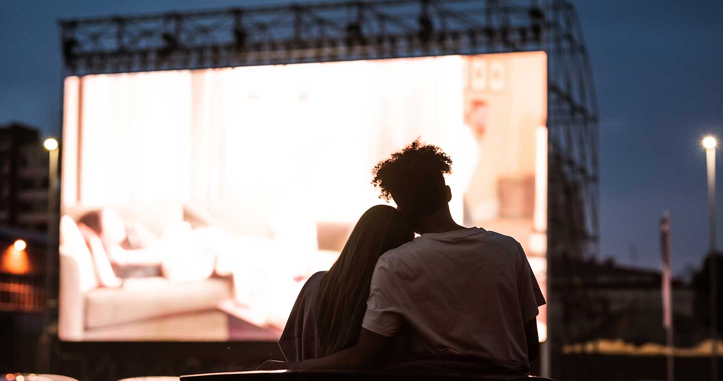 Silhouetted view of attractive young couple, boy and girl embracing, spending time together, sitting in the car while watching a movie in a drive in cinema